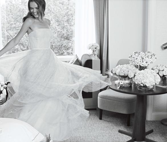 A black and white image of a woman in a white wedding dress spins in a room with bouquets of flowers sitting on table