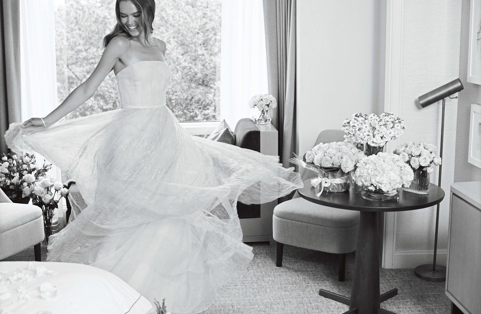 A black and white image of a woman in a white wedding dress spins in a room with bouquets of flowers sitting on table