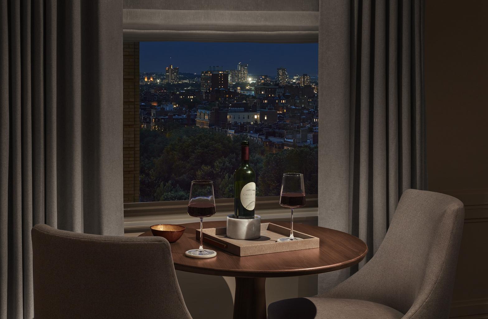 dimly lit sitting area with wine and wine glasses sitting on table beside evening view of city