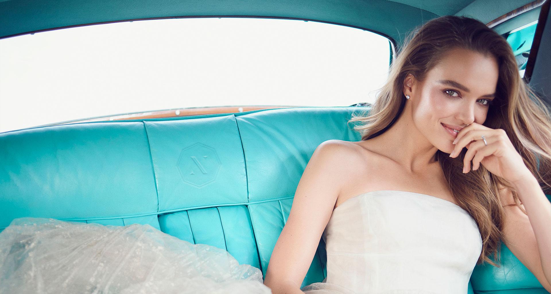 A woman smiling in a white dress in the back seat of car.