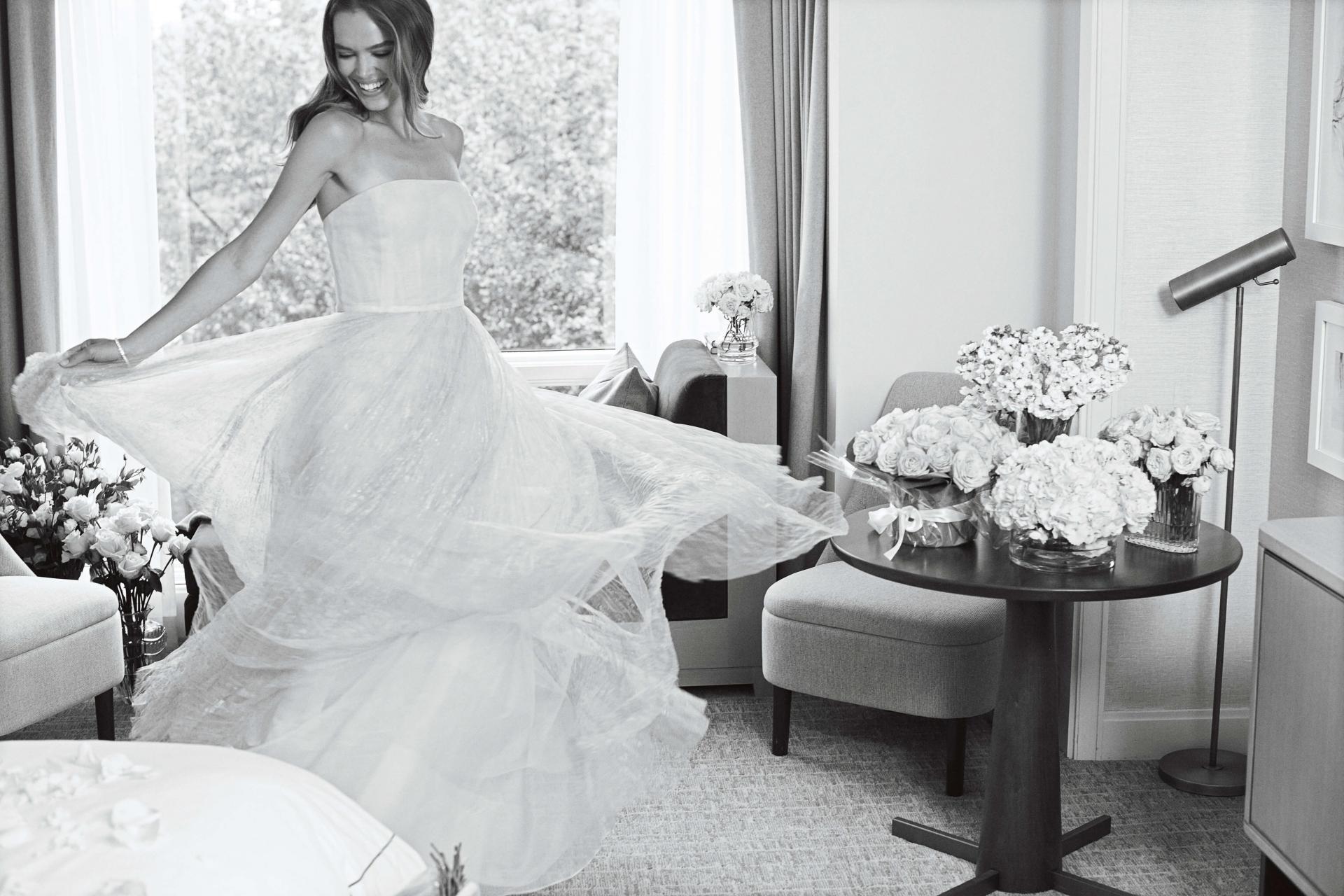 A black and white image of a woman in a white wedding dress spins in a room with bouquets of flowers sitting on table